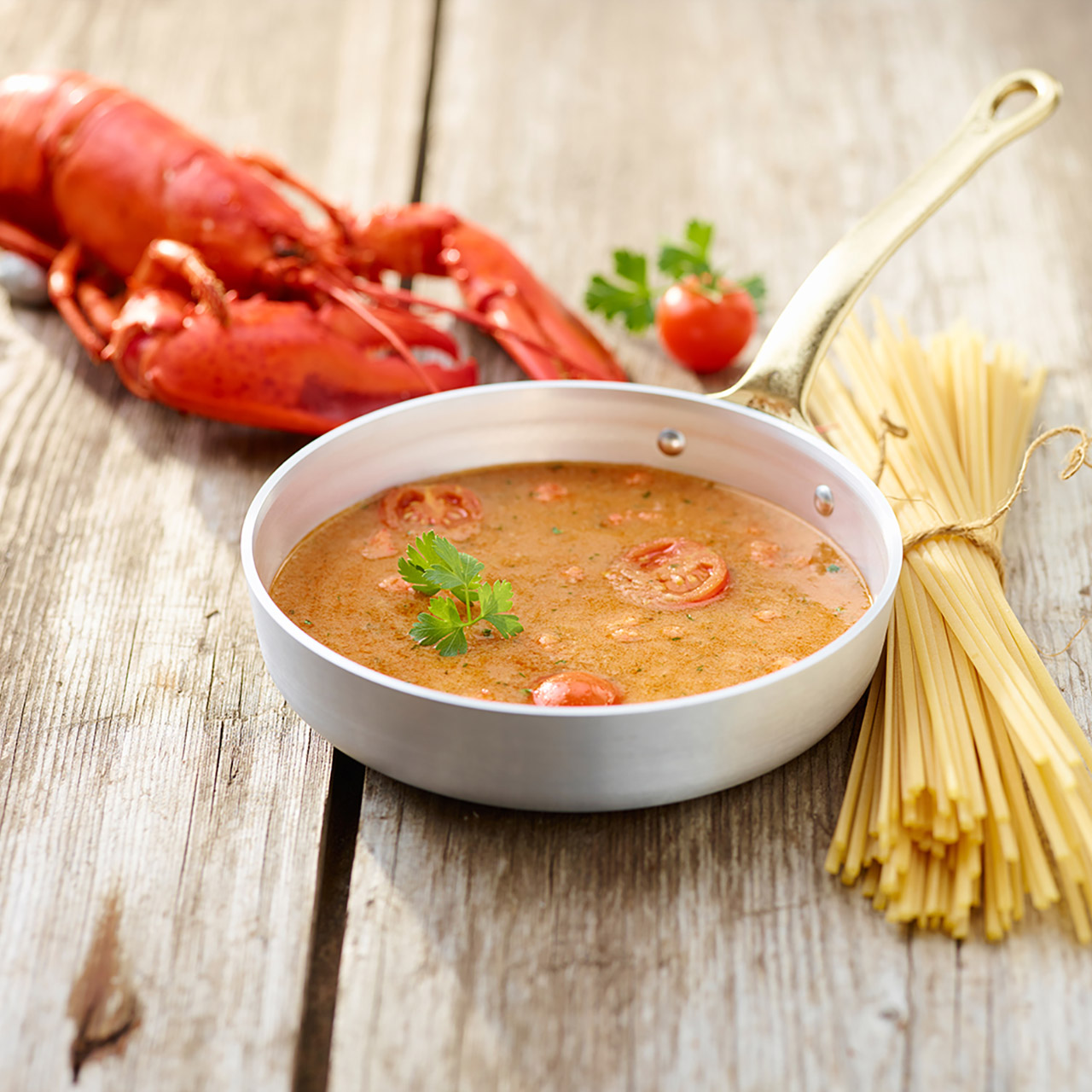 Tegamino contenente una zuppa di pesce targata Arbi, posizionata vicino a un fascio di spaghetti e davanti a un astice e a un pomodorino.